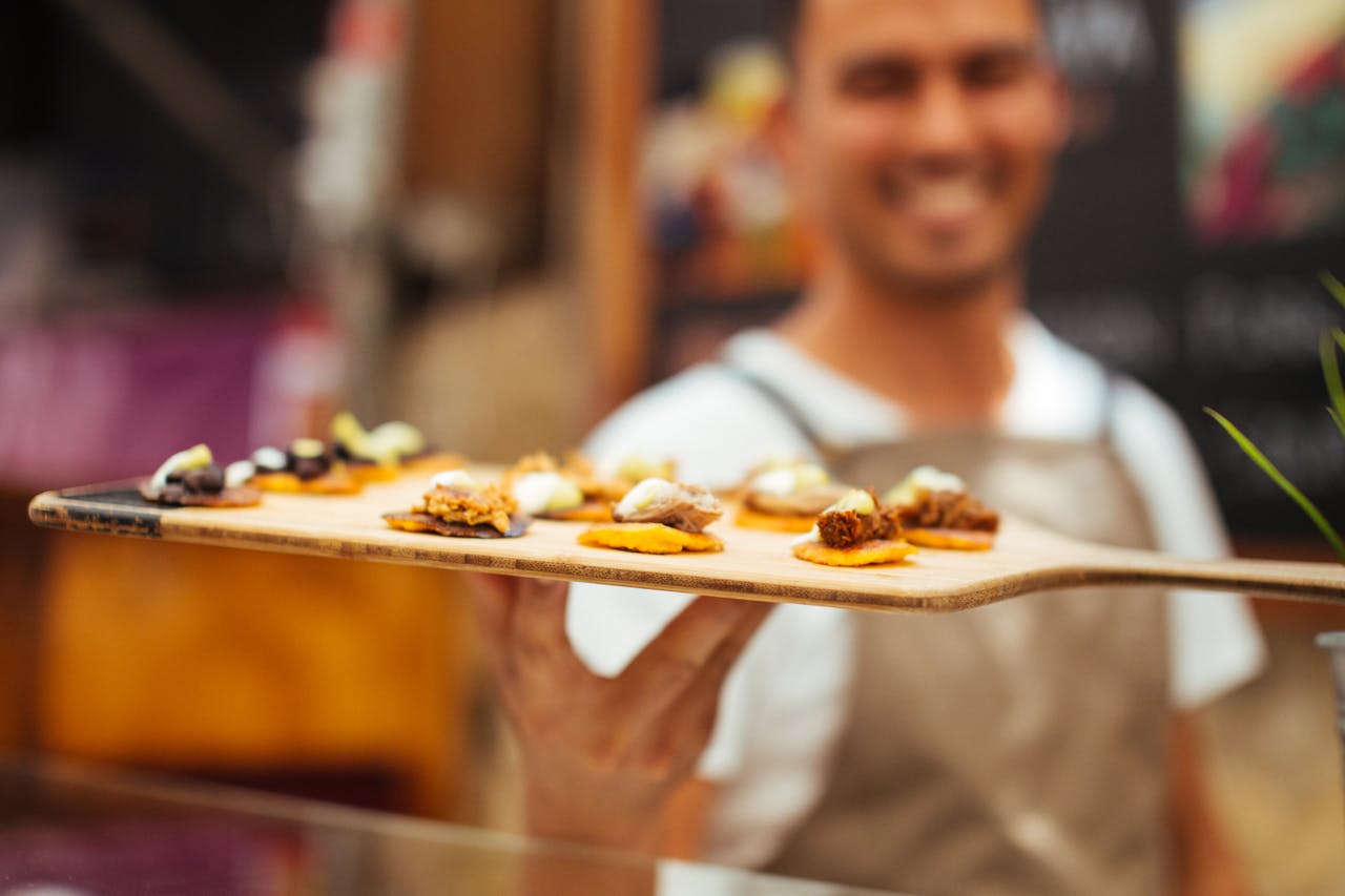 Close-up of a smiling chef presenting a tray of gourmet finger foods in a vibrant market setting.